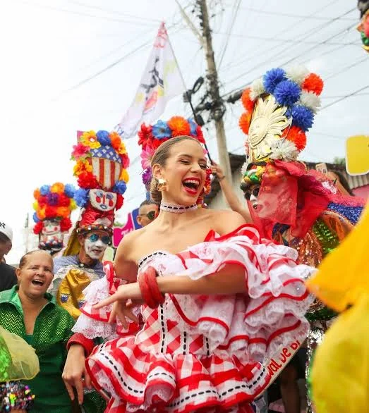 Donna sorridente con un vistoso abito bianco e rosso con volant durante una sfilata del Carnaval de Barranquilla, circondata da costumi colorati e maschere tradizionali.